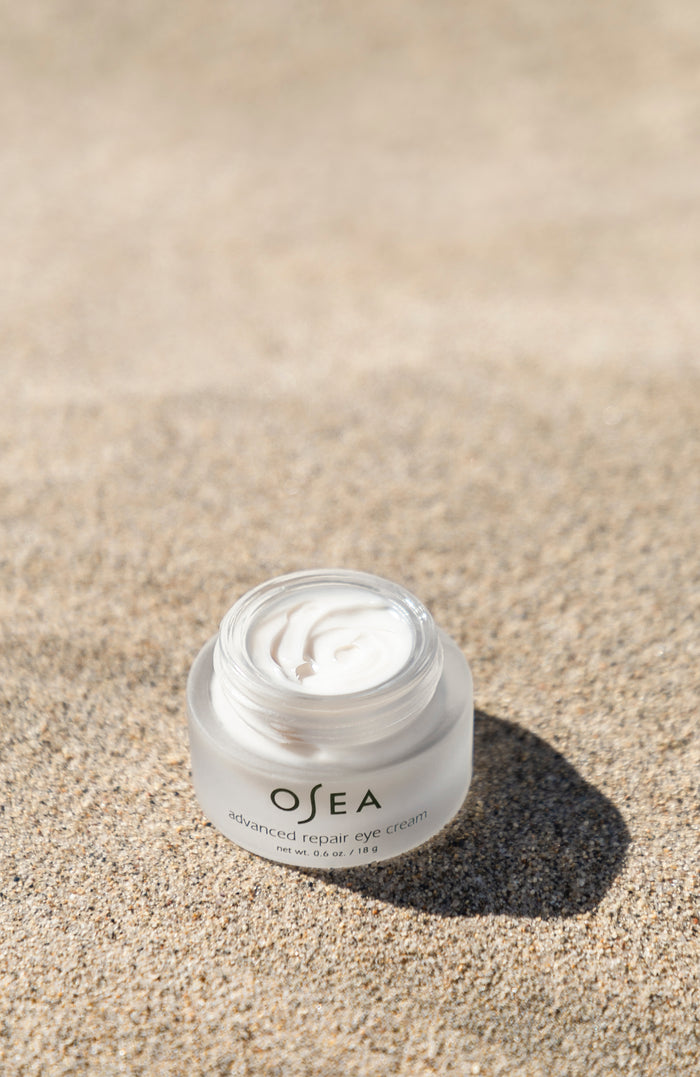 A jar of OSEA Advanced Repair Eye Cream, famed for its anti-aging benefits, rests on the sand. The open lid reveals the white cream inside, casting a shadow alongside sunlit sand textures. The cream is renowned for reducing puffiness.