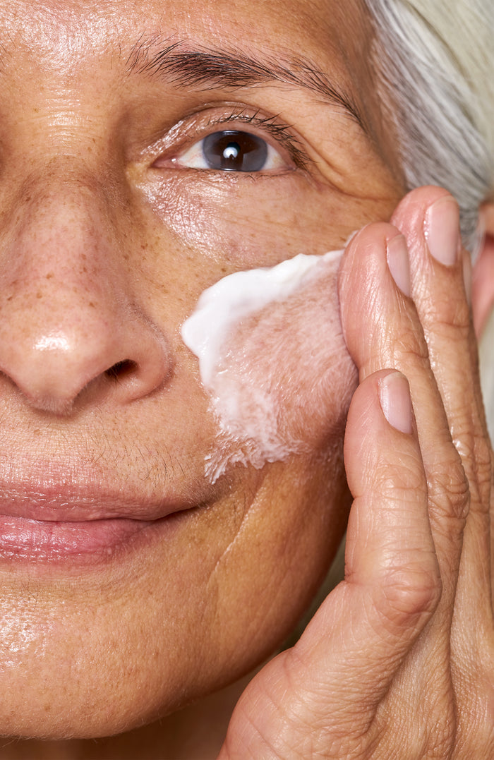 Close-up of a person applying OSEAs Advanced Repair Eye Cream to their cheek. They have light skin, gray hair, and freckles, with a focus on their hand and the creams texture, promising to reduce puffiness while nourishing deeply.