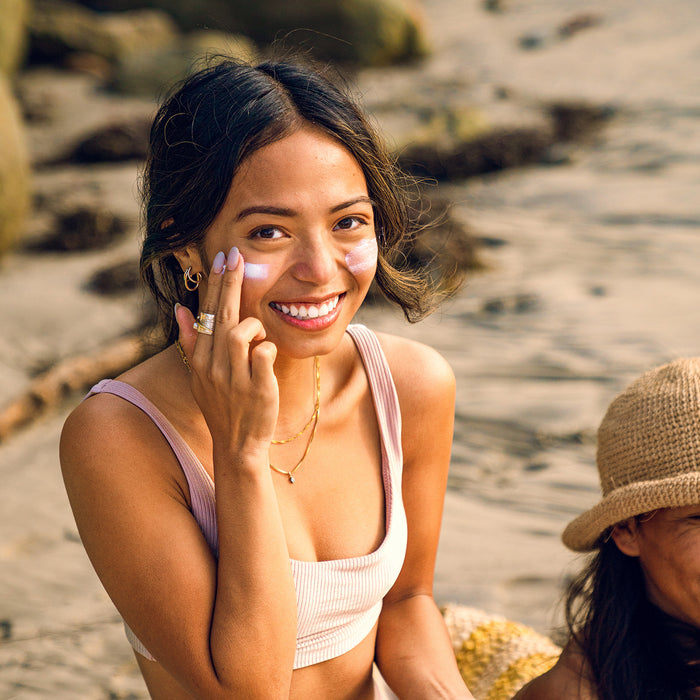 A woman in a light pink top smiles at the camera as she applies Badger Balms Adventure Sport Mineral Sunscreen Cream SPF 50 to her face on a beach, sitting next to someone in a straw hat with sand and rocks in the background.