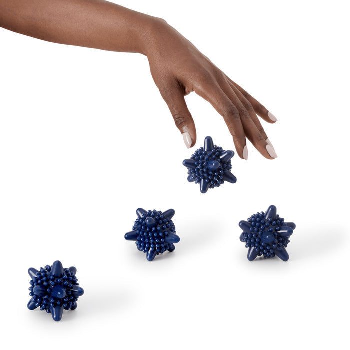 A hand releases a dark blue, spiky Agitator Washer/Dryer Ball by Esembly Baby, crafted from eco-friendly silicone. Three of these identical balls rest on the white surface below, their unique texture and design reminiscent of massage balls.