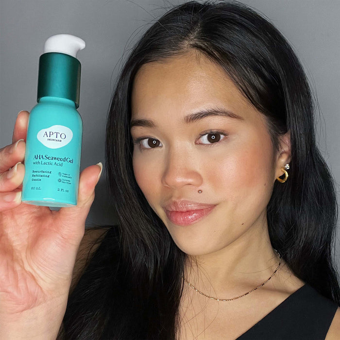 A person with long dark hair holds a bottle of APTO Skincares AHA Seaweed Gel with Lactic Acid, showcasing its resurfacing and deeply exfoliating formula. Smiling in their black top and gold hoop earrings, they stand against a plain gray background.