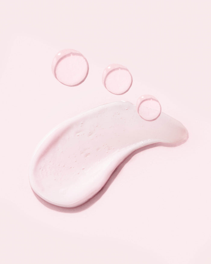 A smear of AHOY LOVE Nourishing Milky Cleanser by Earth Harbor, with Botanic AHAs, is on a pale background. Three small, transparent droplets hover above it, highlighting its lightness and hydrating properties.