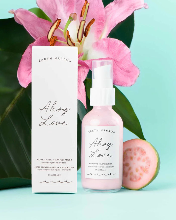 Earth Harbors AHOY LOVE Nourishing Milky Cleanser is shown with its box, nestled among a pink lily, green leaves, and a sliced guava against a light teal backdrop.