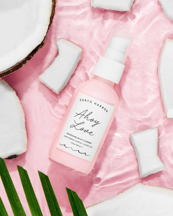 A pink bottle of Earth Harbors AHOY LOVE Nourishing Milky Cleanser sits on a pink marbled surface, surrounded by coconut pieces and greenery.