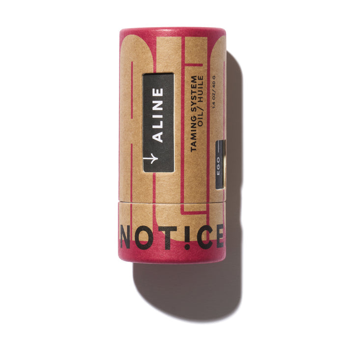 A brown and red cylindrical container displays ALINE Solid Hair Oil by NOT!CE Hair Co. The light enhances its shadowed profile. Designed to tame frizz, its vegan for smooth, luscious hair, with NOTICE written vertically.