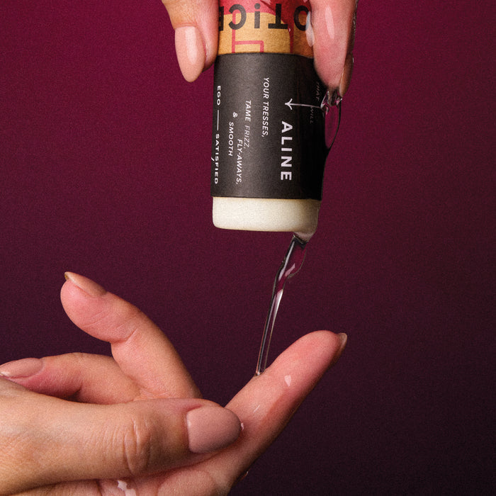 Against a dark red backdrop, a person with manicured nails pours a creamy hair product from a black and beige bottle labeled ALINE Solid Hair Oil by NOT!CE Hair Co, promising to tame frizz effectively with its vegan formula.