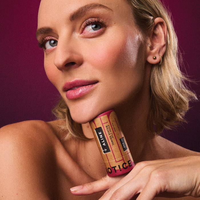 A person with short blonde hair holds the sleek ALINE Solid Hair Oil by NOT!CE Hair Co under their chin, gazing into the distance. The rich magenta background enhances their subtle makeup and highlights this vegan product, perfect for taming frizz.