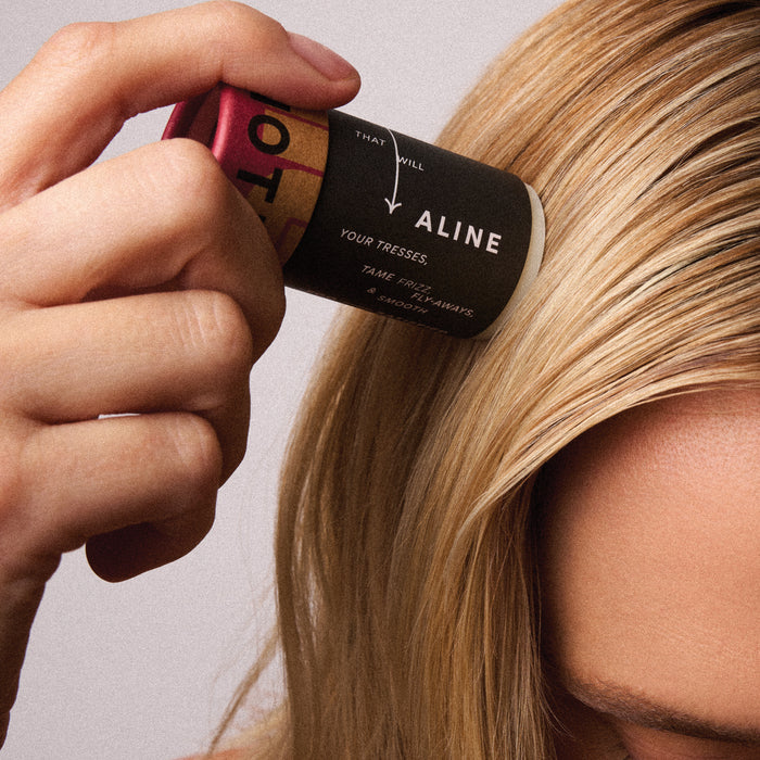 A close-up shows a person using ALINE Solid Hair Oil from NOT!CE Hair Co, applying the vegan-infused product to their blonde hair. Their hand focuses on the scalp near the heads top, reducing frizz, while their face is partially visible.