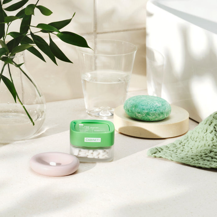 A spa-like bathroom scene with a glass of water, green soap on a pastel dish, a green washcloth, and an All Day Allergy Relief - Cetirizine HCl 10 mg container by Cabinet Health enchants the light counter. A vase with leafy stems enhances the calming atmosphere.