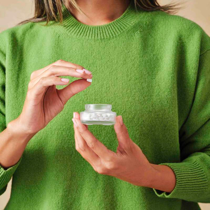 Wearing a green sweater, an individual carefully lifts cream from a jar, much like handling Cabinet Healths All Day Allergy Relief - Cetirizine HCl 10 mg. The soft beige background amplifies the calming effect of this sustainable medicine.
