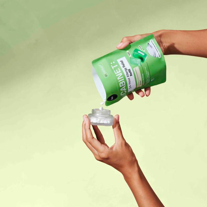 Hands pour white tablets from a green pouch marked Cabinet Health: All Day Allergy Relief - Cetirizine HCl 10 mg into a transparent container on a light green background.