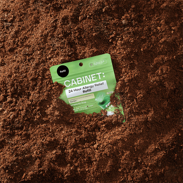 A green All Day Allergy Relief - Cetirizine HCl 10 mg packet by Cabinet Health peeks from brown soil, highlighting sustainable all-day allergy relief for health and planet.
