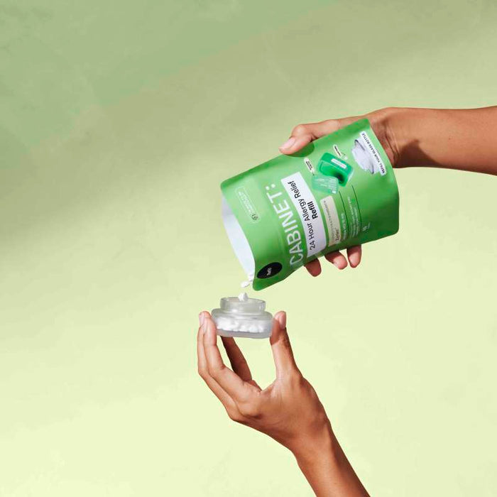 Two hands hold a green refill pack labeled CABINET: All Day Allergy Relief Refill - Cetirizine HCl 10 mg. One hand pours the contents into a clear bottle with a white cap against a light green background, offering effective allergy relief similar to Zyrtec.