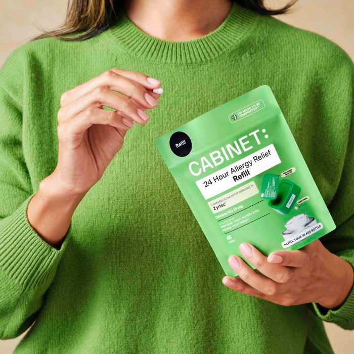 A person in a green sweater holds a packet labeled All Day Allergy Relief Refill - Cetirizine HCl 10 mg by Cabinet Health, comparable to Zyrtec. The packet shows images of the refill container and tablets, offering cetirizine HCl-based relief for allergies.