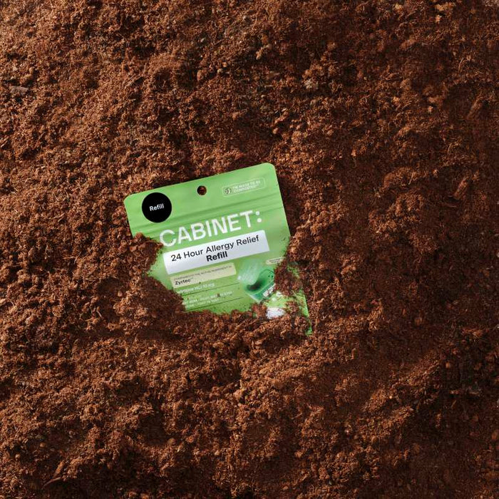A green package of Cabinet Healths All Day Allergy Relief Refill, featuring cetirizine HCl 10 mg and comparable to Zyrtec, is partially buried in rough brown soil while uprightly displaying its branding and text.