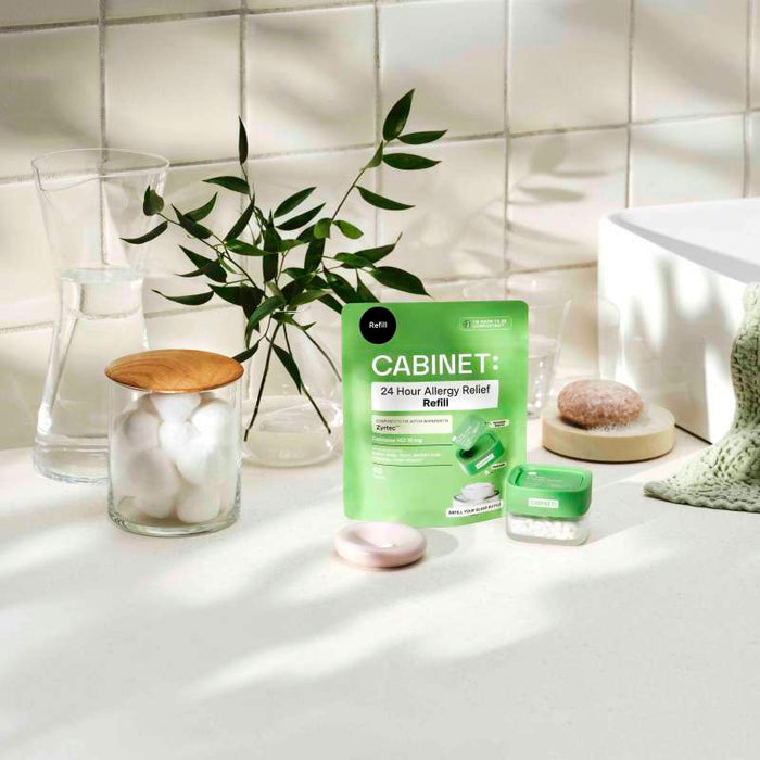 A bathroom countertop features a green pouch labeled Cabinet Health: All Day Allergy Relief Refill - Cetirizine HCl 10 mg, similar to Zyrtec, next to a glass jar of cotton balls, a small green container, and a clear glass pitcher. Green leafy branches in a vase grace the scene as soft light softly illuminates.