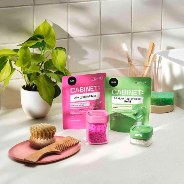 On a bathroom countertop sits pink and green packaged Cabinet Health All Day Allergy Relief Refill tablets containing cetirizine HCl for Zyrtec-like effectiveness. Nearby are a white potted plant, wooden comb, brush, toothbrushes in a holder, and green sponge by the sink.