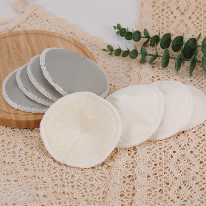 Haakaas All-Day Reusable Nursing Pads, in gray and beige, are showcased on lace cloth. Some are neatly stacked on a round wooden tray beside a sprig of green leaves.