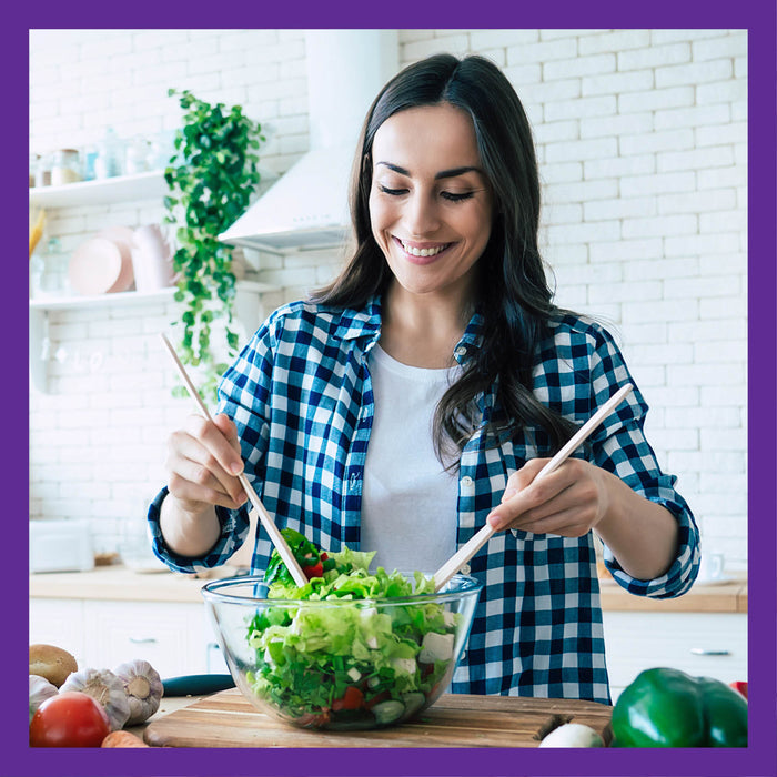 In a kitchen with white brick walls, shelves of plants, and colorful countertop vegetables, a woman in a blue checkered shirt smiles as she uses salad tongs to prepare a salad in a clear bowl. Shes mindful of digestive health with New Chapters All Flora Probiotic + Prebiotic.