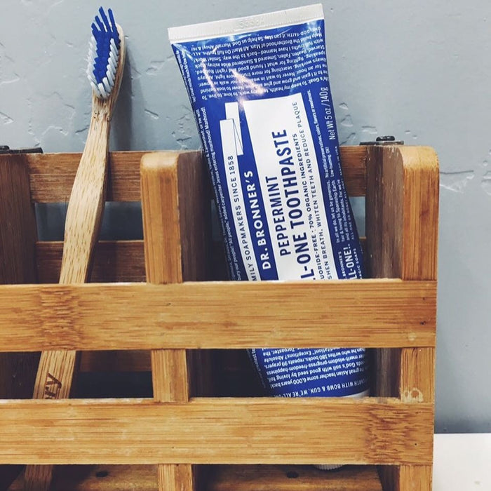 A wooden holder holds a blue toothbrush and a tube of Dr. Bronners All-One Toothpaste, fluoride-free, set against a gray wall.