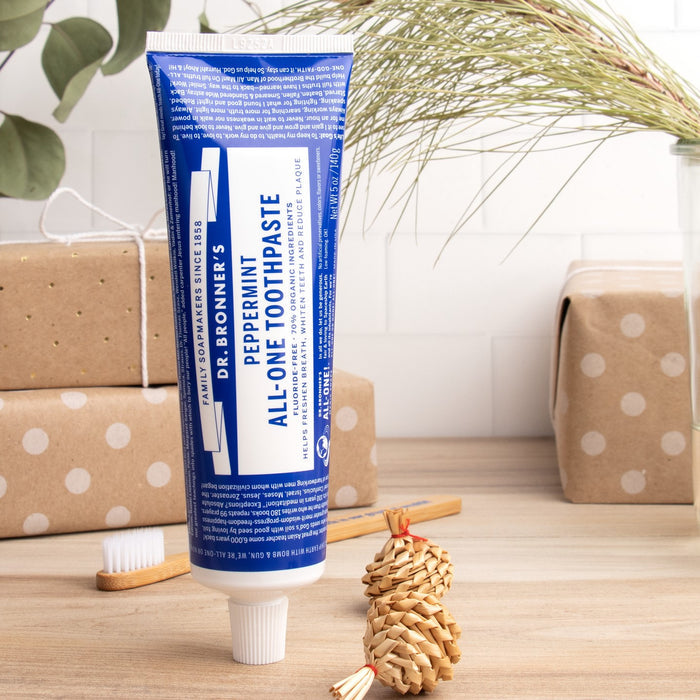 A tube of Dr. Bronners All-One Toothpaste stands upright on a wooden surface, surrounded by a toothbrush, wrapped gifts, dried plants, and a braided straw ornament.