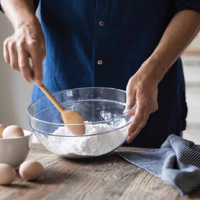 A person in a blue shirt mixes flour in a transparent bowl using a Bambu Mixing Spoon. A white bowl with eggs sits on the rustic wooden table next to a folded blue cloth, creating an eco-friendly kitchen scene.