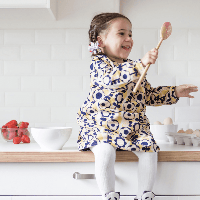 A young girl in a floral dress sits on the kitchen counter, smiling with a Bambu Mixing Spoon in hand. Nearby are a bowl, strawberries, and a carton of eggs. The kitchen has an eco-friendly vibe with its white tiled backsplash.