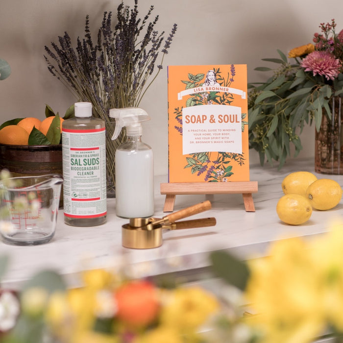 A kitchen counter displays Dr. Bronners All-Purpose Cleaner, a spray bottle, measuring cups, lemons, the book Soap & Soul on a stand, plus vases with flowers and greenery in the background.