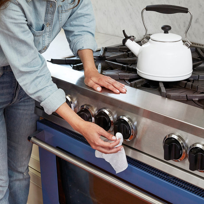 Wearing blue jeans and a light denim shirt, a person uses method All-Purpose Compostable Cleaning Wipes to clean the stainless steel stovetop, focusing on the control knobs. A white kettle sits on a burner as every surface shines against the blue oven door.
