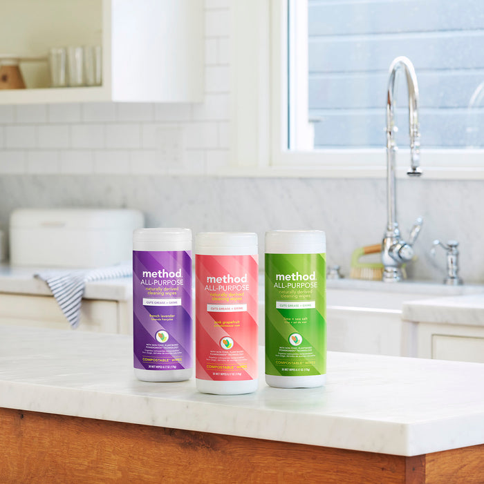 Three containers of methods All-Purpose Compostable Cleaning Wipes sit on a kitchen countertop, their vibrant designs in purple, pink, and green shining in the sunlight, showcasing their natural cleaning power for various surfaces.