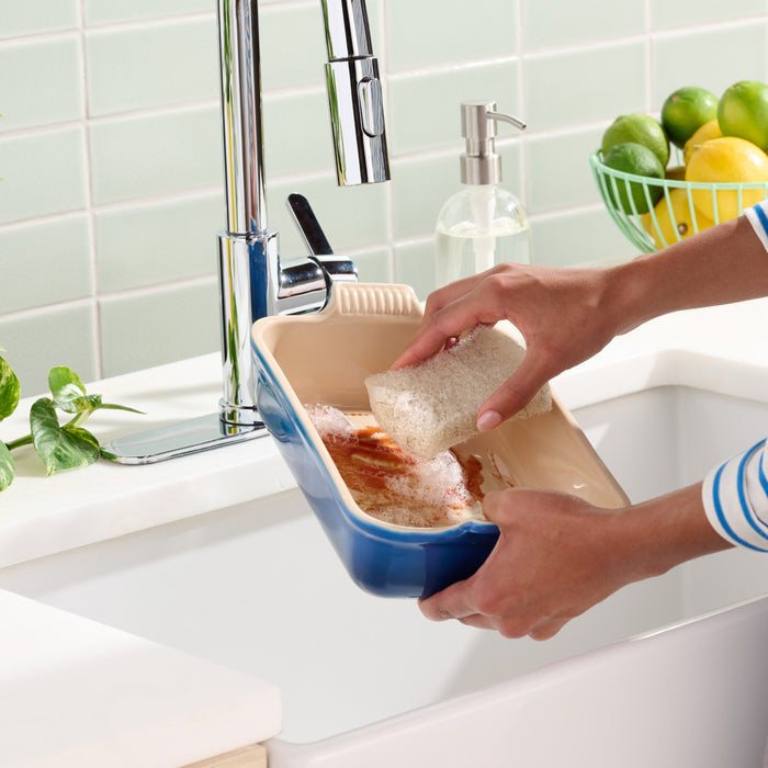 A person cleans a stained ceramic baking dish at the kitchen sink using Grove Co. All-Purpose Coconut Scouring Pads; a soap dispenser and a bowl of limes and lemons are seen in the background.