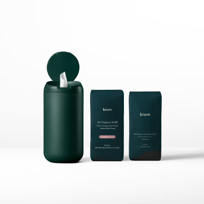 An open dark green cylinder displays naturally derived cleaning wipes next to two refill packs titled biom All-Purpose Wipe Refills. The minimalist design of these eco-friendly products is highlighted by a plain white background.
