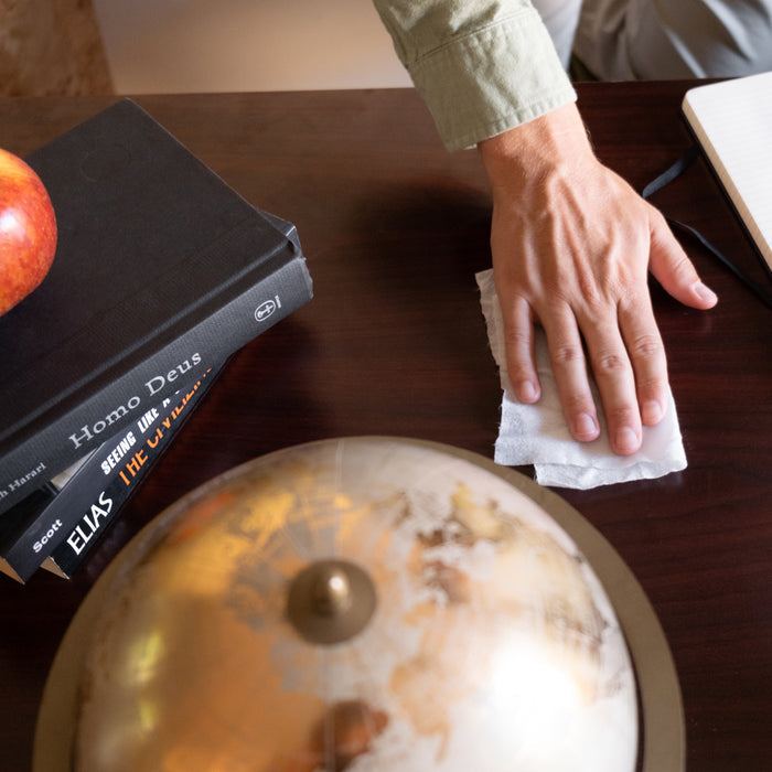 A person uses Biom All-Purpose Wipe Refills to clean a wooden table. On the table sit a globe, two stacked books titled Homo Deus and EIMAS, an apple, and a corner of an open notebook in view.