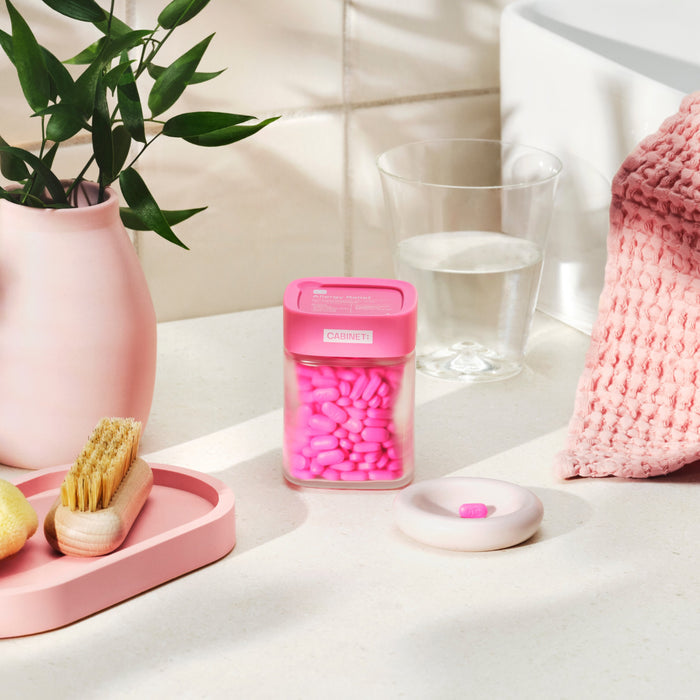 On a bathroom counter, a pink jar of Cabinet Healths Allergy Relief - Diphenhydramine 25 mg stands next to a water glass, vase with leaves, and pink tray with nail brush. Nearby, a pill rests on a dish beside a pink towel. The bright setting feels calm and organized.