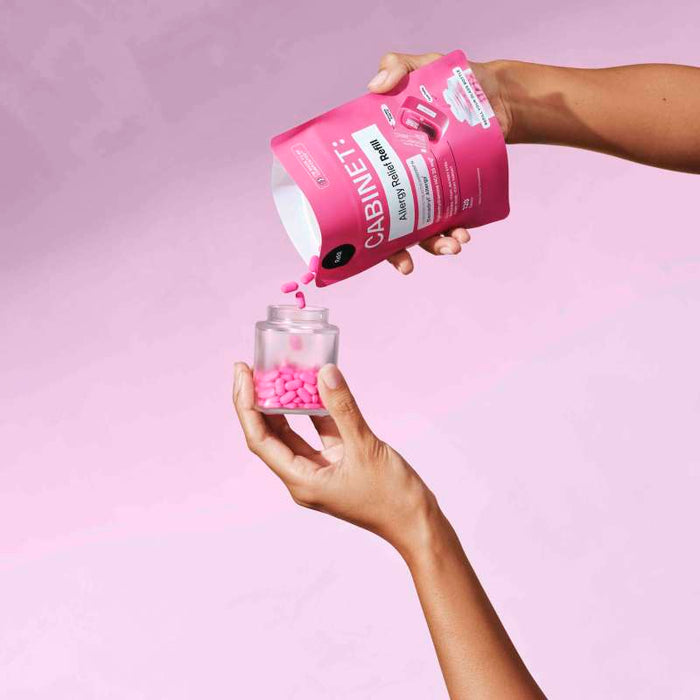 The image shows hands transferring pink pills from a Cabinet Health packet labeled Allergy Relief - Diphenhydramine 25 mg into a clear jar against a light pink gradient background, highlighting sustainable allergy relief.