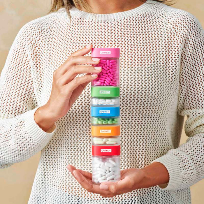 A person wearing a white knit sweater holds a vertical stack of five transparent containers with pink, blue, green, red, and white pills. Each has a colored lid and is labeled as Allergy Relief - Diphenhydramine 25 mg by Cabinet Health, promoting sustainable medicine solutions.