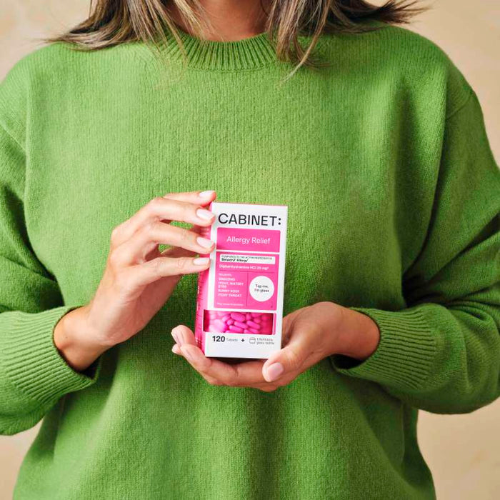 A person in a green sweater holds a box of Cabinet Healths Allergy Relief - Diphenhydramine 25 mg, showcasing its sustainable medicine approach. The pink and white package offers non-drowsy antihistamine relief for allergy symptoms, containing 120 tablets.