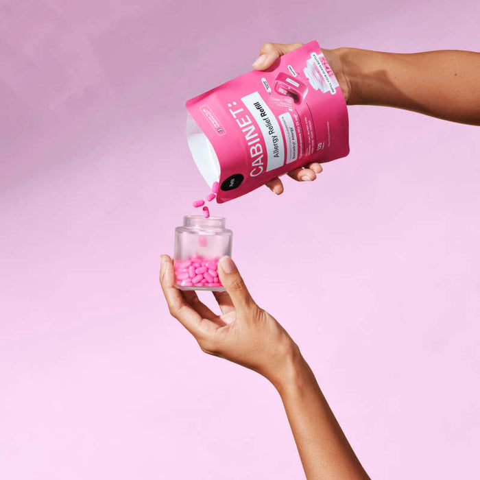 Two hands against a pink background: one pours Diphenhydramine 25 mg pills into a jar from a pink Allergy Relief Refill package by Cabinet Health with sustainable, compostable materials.