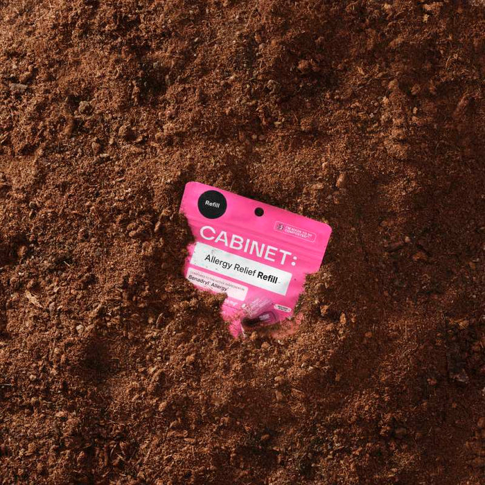A compostable pink packet labeled Cabinet Health: Allergy Relief Refill - Diphenhydramine 25 mg with a durable, resealable design is partially buried in soil, standing out against the brown earth.