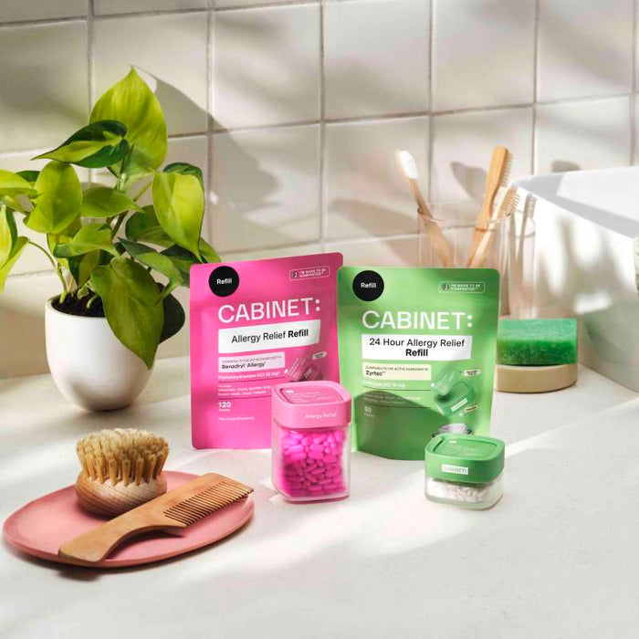 Bathroom countertop showcasing Cabinet Healths compostable-packaged Allergy Relief Refill - Diphenhydramine 25 mg, including pink and green refill pouches and small containers. Accompanied by a plant, brush, comb, and soap dish against a white tiled background.