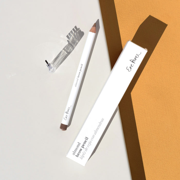 An Ere Perez Almond Brow Pencil with a clear cap lies on its white box against overlapping white and light brown paper, its shadows enhancing the nourishing almond oil infused within.