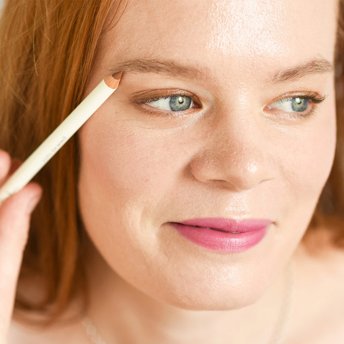 With light brown hair, blue-eyed and subtly smiling while wearing pink lipstick, a person uses the Ere Perez Almond Brow Pencil on their right eyebrow. Their smooth complexion is enhanced by nourishing almond oil against a softly blurred background.