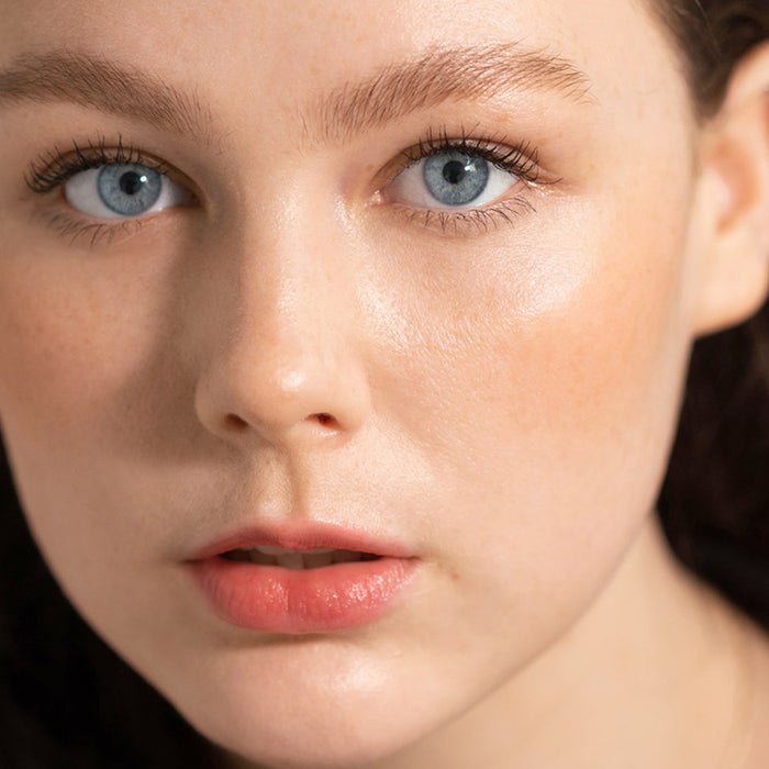 Close-up of a fair-skinned person with blue eyes and rosy lips. They have natural makeup, enhanced eyelashes, and light freckles accented by almond oil. The look is perfected with gentle strokes from Ere Perezs Almond Brow Pencil as they gaze directly at the camera.
