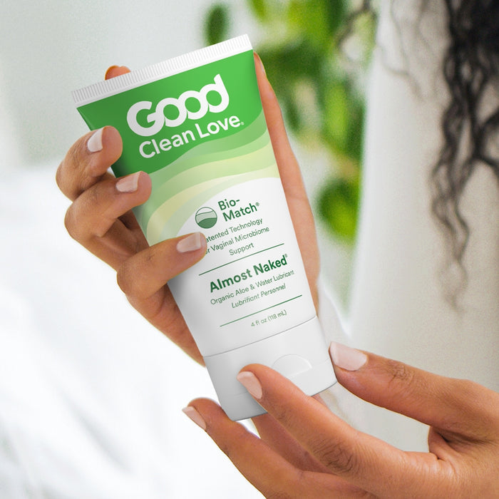 A person with light skin and well-manicured nails holds a green and white tube of Good Clean Loves Almost Naked Organic Personal Lubricant, featuring Bio-Match technology and a water-based formula for vaginal health.