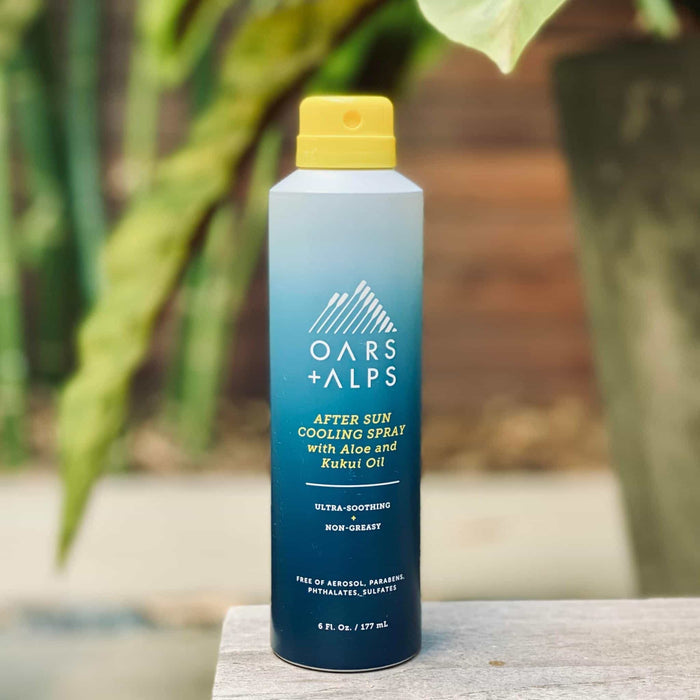 A can of Oars + Alps Aloe Cooling Spray rests outdoors on a surface, surrounded by green leaves and a wooden backdrop.