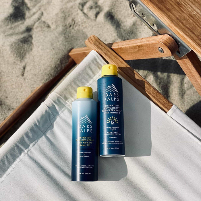 Two Oars + Alps Aloe Cooling Spray bottles with yellow caps sit on a white beach chair. Sand and the wooden frame are seen behind them.