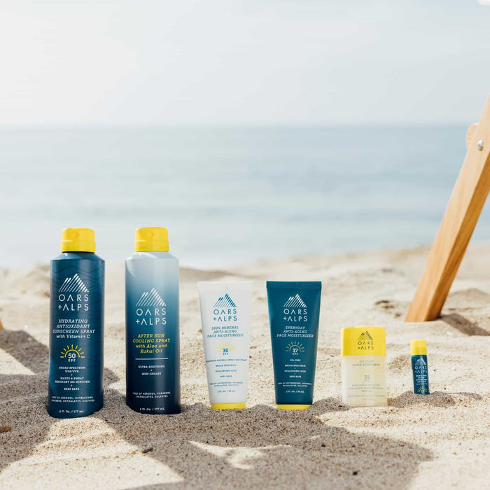 Oars + Alps Aloe Cooling Spray, rich in antioxidants and Aloe Vera, stands upright on sandy beach with the ocean and a wooden beach chair in the bright sunlight.