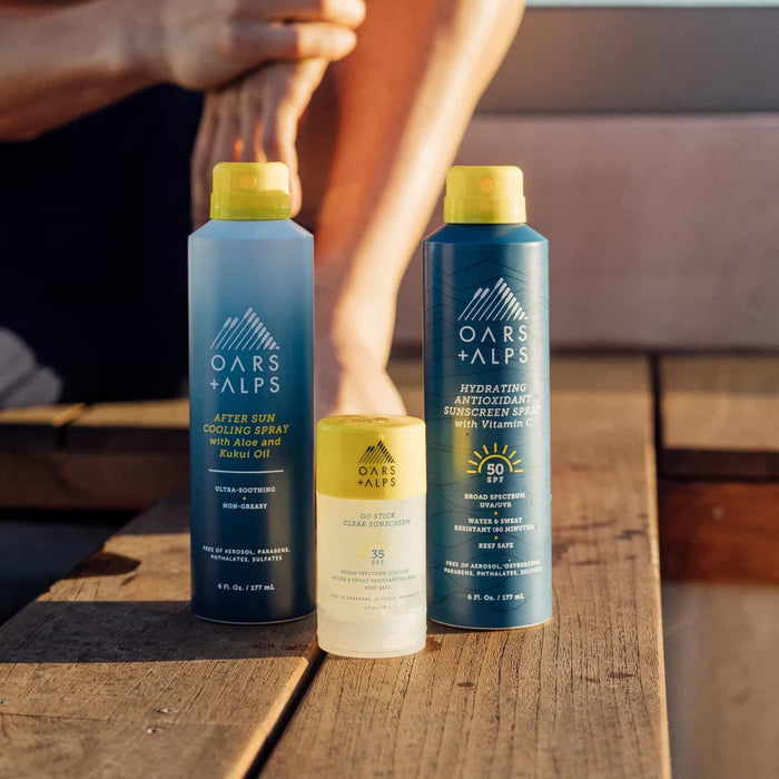 Three Oars + Alps skincare products—Aloe Cooling Spray, Hydrating Antioxidant Sunscreen Spray, and Go Stick Clear Sunscreen—are displayed on a wooden surface in sunlight with a person visible in the background.