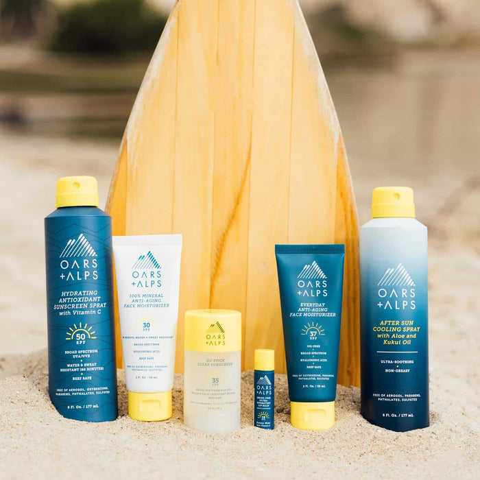 Oars + Alps Aloe Cooling Spray, along with other skincare and sun protection products, stands upright on sand before a wooden surfboard at the beach.