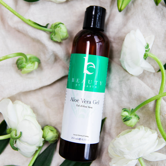 Beauty by Earths Aloe Vera Gel, with its green label, sits gracefully on beige fabric surrounded by white flowers and green leaves, creating a natural, refreshing aesthetic ideal for those seeking pure skincare.